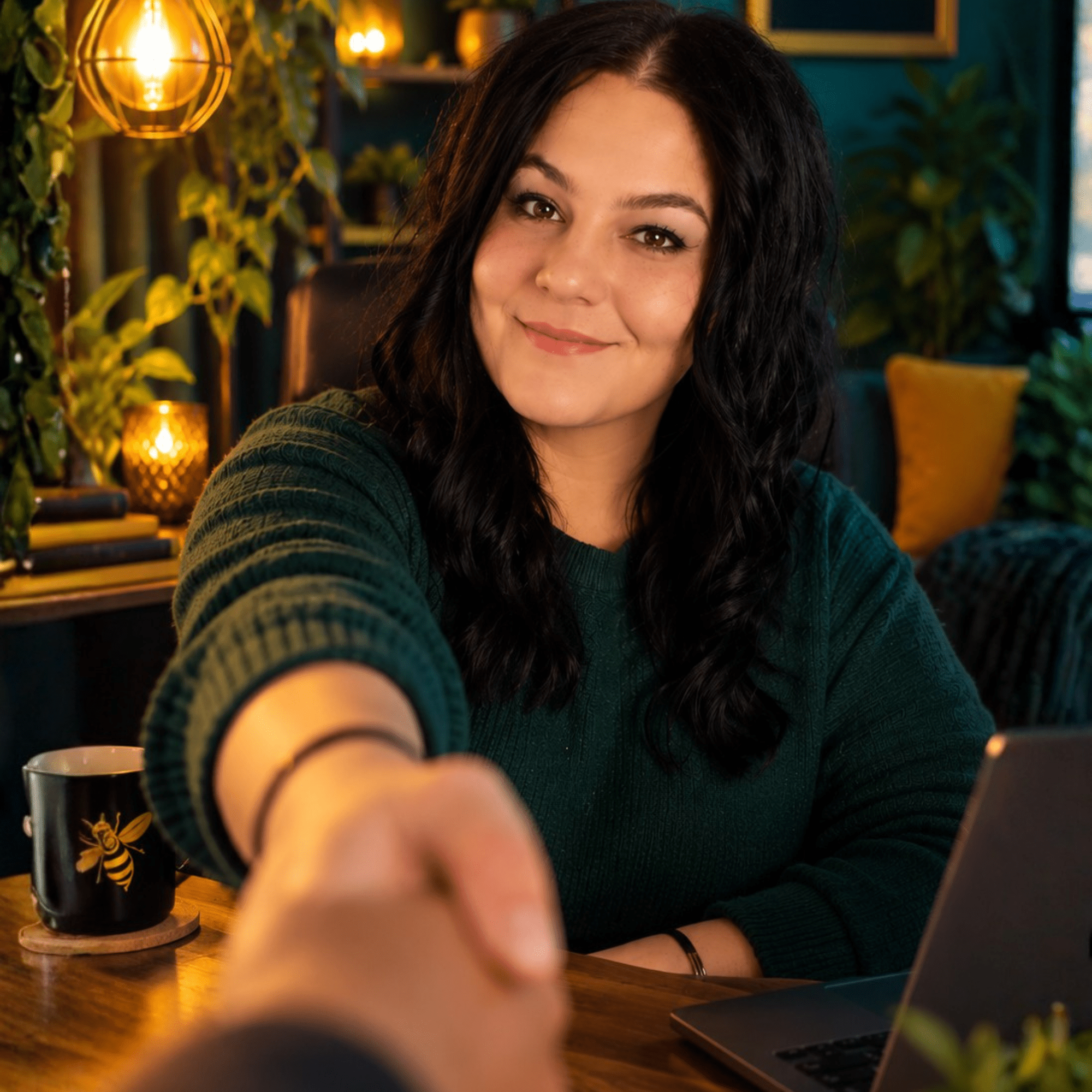 Friendly female business owner smiling warmly at the camera and reaching out for a handshake in a cozy teal and yellow creative office.