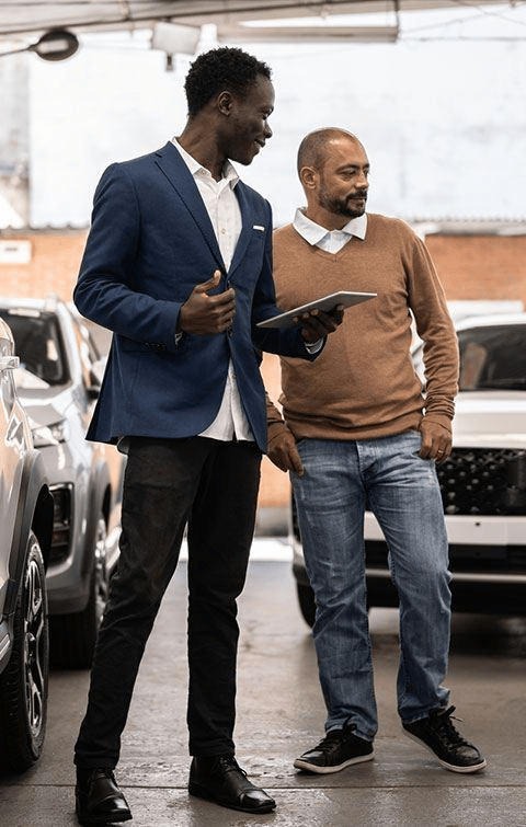 A realistic car dealership scene with two salespeople: one extremely polished and traditionally “salesy,” and another dressed casually and approachable. This directly illustrates the analogy in the blog.